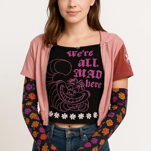 May include: A woman wearing a black crop top with the text "We're all MAD here" and a Cheshire Cat graphic. She is also wearing a pink zip-up jacket, blue jeans, and black arm sleeves with a rose pattern.