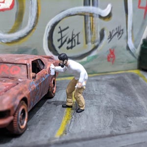 May include: A miniature scene depicts a rusty brown car with the word "NITRO" in red on the windshield. A small figure in a white shirt and tan pants appears to be spray painting the car. The background features graffiti art and a green dumpster.