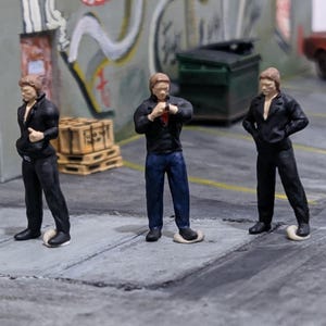 Puede incluir: Figuras en miniatura de tres personas con chaquetas y pantalones negros, de pie sobre una superficie gris. Una figura lleva vaqueros azules. La escena incluye un edificio, graffiti, un contenedor de basura, un coche y palets de madera.