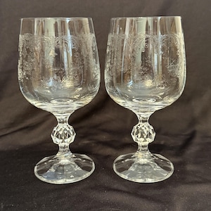 May include: Two clear crystal wine glasses with etched floral designs. Each glass has a diamond-shaped stem and a round base. The glasses are displayed on a dark background.