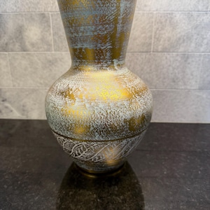 May include: A decorative vase with a bulbous base and a flared top. The vase has a gold metallic finish with a distressed, textured appearance. The gold is accented with patches of white and light blue. The base features a white, etched leaf design.