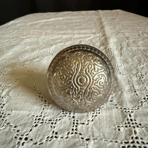 May include: A round, silver-toned metal door knob featuring an intricate, embossed scrollwork pattern across its surface. The knob has a central circular detail surrounded by symmetrical, swirling decorative motifs in a metallic finish.