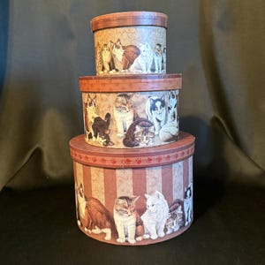 May include: A set of three round, stacked cardboard storage boxes featuring a repeating pattern of various cats in shades of brown, white, and gray. Each box lid has a brown border with a small red paw print design around the circumference.