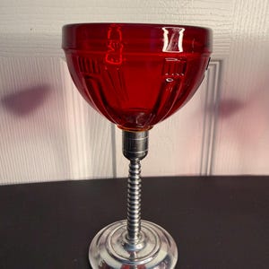 May include: A vintage red glass lamp shade with a ribbed design, mounted on a silver-colored metal base. The base has a round, decorative foot and a flexible, coiled neck. The lamp is likely an antique or retro decorative item.