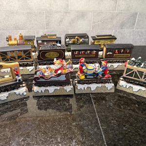 May include: A collection of Christmas train cars and decorative figurines. The train cars are brown, gold, and black, with various designs. Santa Claus in a sleigh, elves, and a display with the year 1998 are featured. The figurines sit on a snowy base.