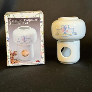 May include: A white ceramic potpourri simmer pot with a two-piece design, featuring a graphic of two swans facing each other with a pink heart. The box displays the text Ceramic Potpourri Simmer Pot and notes that potpourri and candle are not included.