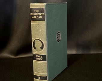 May include: A hardcover book titled The Innocents Abroad by Mark Twain with a teal green cloth cover. The spine features black panels with white text, a silhouette portrait, and embossed scrollwork patterns. A circular emblem is on the front cover.