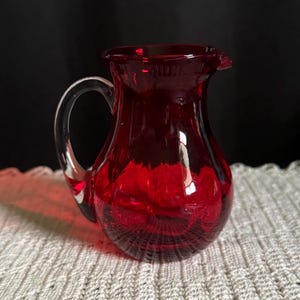 May include: A ruby red glass pitcher with a clear handle and spout. The pitcher is sitting on a white, textured surface. The pitcher is likely used for serving beverages.