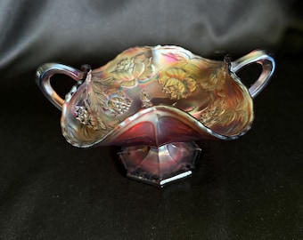May include: A footed carnival glass bowl with two handles, featuring an iridescent purple and red finish with a molded floral pattern. The bowl has a scalloped rim and sits on a hexagonal base, displaying a reflective surface with metallic highlights.