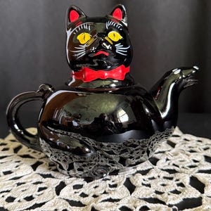May include: A black ceramic teapot shaped like a cat, with yellow eyes, red accents, and a red bow tie. The teapot sits on a white lace doily, with a dark background.