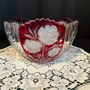 May include: A clear glass bowl with a ruby red interior and frosted white rose designs. The bowl has a scalloped rim and vertical grooves, creating a textured appearance. This decorative bowl is ideal for serving or display.