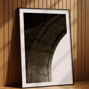 Può includere: Fotografia in bianco e nero incorniciata di una struttura ad arco in pietra. L'immagine è appoggiata a una parete e un pavimento in legno, con ombre della luce solare. L'arco è fatto di mattoni usurati.