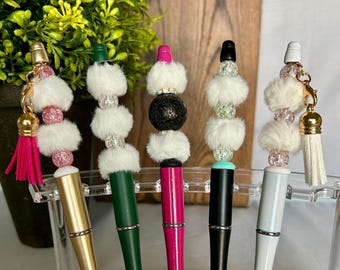 Op de afbeelding: Vijf decoratieve pennen met verschillende kleuren en versieringen. Elke pen heeft een ander gekleurde schacht, waaronder goud, groen, roze, zwart en wit. Ze zijn versierd met witte imitatiebont, kralen en kwastjes, wat zorgt voor een uniek en stijlvol ontwerp.