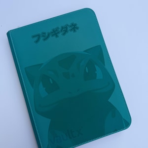 Peut inclure: Classeur de cartes de couleur teal avec un motif Bulbasaur et du texte japonais. Le classeur a une bordure cousue et le logo VaultX. Le motif est d'une nuance de teal plus foncée.