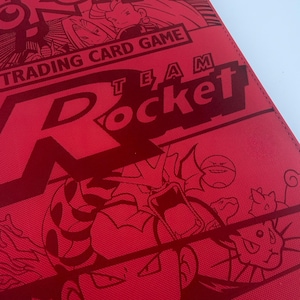 May include: A red Pokémon Trading Card Game Team Rocket binder featuring a textured surface with dark red line art illustrations of characters and creatures, including the text Pokémon Trading Card Game and Team Rocket printed in bold lettering.