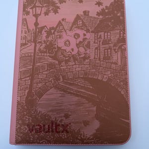 Puede incluir: Un archivador de tarjetas rosa con una ilustración marrón de una escena de pueblo, que incluye un puente, casas y una farola. La palabra "vaultx" está impresa en la esquina inferior izquierda. El archivador está diseñado para guardar tarjetas.