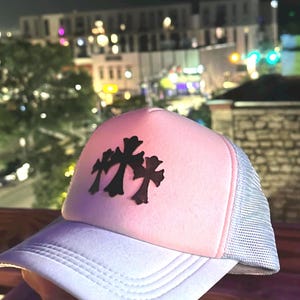 May include: Pink trucker hat with a white brim and mesh back. The hat features a black cross design on the front. The hat is worn outdoors at night with city lights in the background.