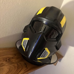 May include: Black helmet with yellow stripes and accents. The helmet has a visor and is resting on a reflective surface. The design is futuristic and sleek, suggesting a cosplay or costume item.