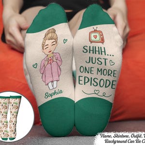 May include: A pair of cream-coloured socks with dark green cuffs and toes. One sock features an illustration of a person with a bun eating popcorn and the name Sophia. The other sock displays a television icon and the text SHHH... JUST ONE MORE EPISODE.