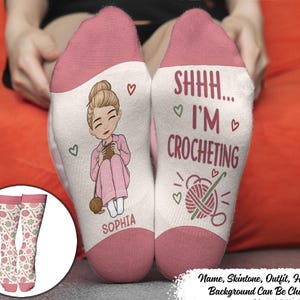 May include: A pair of white socks with pink cuffs and toes. One sock features a cartoon illustration of a person with a bun hairstyle crocheting, with the name SOPHIA printed below. The other sock displays the text SHHH... I'M CROCHETING and a yarn ball.