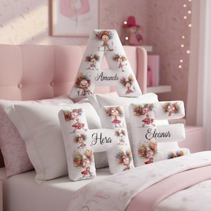 May include: White decorative pillows shaped like the letters A, H, and E, with the names "Amanda", "Hera", and "Eleanor" printed on them. Each pillow features a colorful cartoon illustration of a girl. The pillows are on a pink bed.