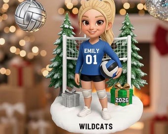 Peut inclure: Une décoration de Noël suspendue représentant une joueuse de volley-ball blonde en maillot bleu avec le nom EMILY et le numéro 01, tenant un ballon. La base blanche porte le texte WILDCATS, deux sapins verts et un cadeau marqué 2026.