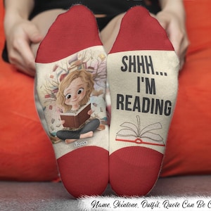 Op de afbeelding: Een paar crèmekleurige sokken met rode tenen en hielen. De linkersok toont een illustratie van een persoon die een boek leest, omringd door zwevende boeken. De rechtersok toont de zwarte tekst SHHH... I'M READING boven een icoon van een boek.