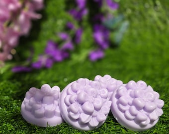 May include: Four round, lavender-coloured soaps with a floral design are arranged on a bed of green grass. The soaps have a detailed flower pattern on their surface. Blurred purple flowers in the background add a soft, natural touch.