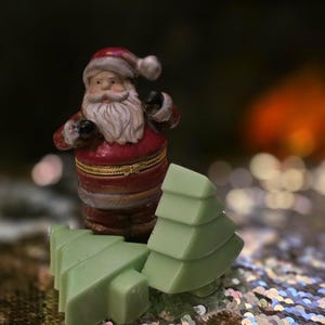 Op de afbeelding: Een kleine kerstmanfiguur in een rood en wit kostuum met een lange witte baard staat naast twee groene kerstboomversieringen. De achtergrond is wazig met bokeh-lichten, wat een feestelijke vakantiescène suggereert.
