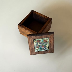 Può includere: Scatola di legno quadrata aperta con coperchio. Il coperchio presenta un design floreale quadrato e colorato con accenti rossi, blu e verdi. La scatola è in legno scuro.