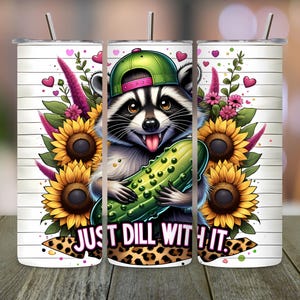 May include: A white tumbler with a cartoon raccoon wearing a green cap and holding a pickle. The tumbler is decorated with sunflowers, pink hearts, and the phrase "JUST DILL WITH IT" in a playful font.