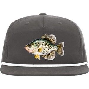 May include: A dark gray cap featuring a detailed illustration of a crappie fish. The fish is depicted with a mix of green, yellow, and black markings. A white rope detail accents the brim of the cap, offering a classic, outdoorsy aesthetic.