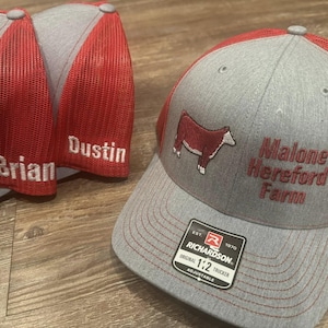 May include: Grey and red trucker caps with embroidered text and a cow graphic. One cap says "Malone Hereford Farm." Two other caps have the names "Brian" and "Dustin" embroidered. The caps have an adjustable strap.
