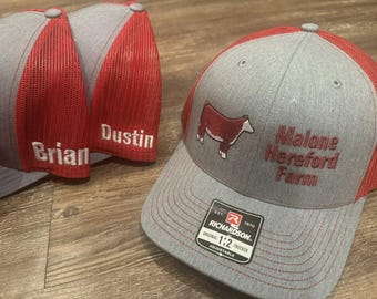 Op de afbeelding: Grijze en rode truckerpetten met geborduurde tekst en een koeienafbeelding. Eén pet zegt "Malone Hereford Farm". Twee andere petten hebben de namen "Brian" en "Dustin" geborduurd. De petten hebben een verstelbare band.