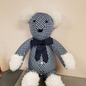 May include: A blue and white gingham teddy bear with white fur accents. The bear is wearing a navy blue bow tie and has black button eyes. The bear is sitting on a light gray surface.