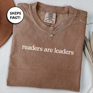 Könnte beinhalten: Ein braunes T-Shirt mit dem weißen Aufdruck "readers are leaders". Eine silberne Halskette mit einem kleinen Anhänger liegt auf dem Shirt. Das Shirt hat ein Comfort Colors-Etikett.