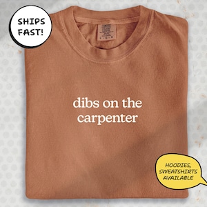 a brown t-shirt with the text "dibs on the carpenter" printed on it. The t-shirt is folded and displayed on a surface, with a speech bubble indicating that it is available for shipping.