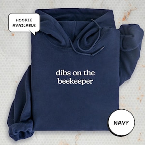 A navy blue hoodie with the text "dibs on the beekeeper" printed on it, along with a speech bubble indicating that the hoodie is available.