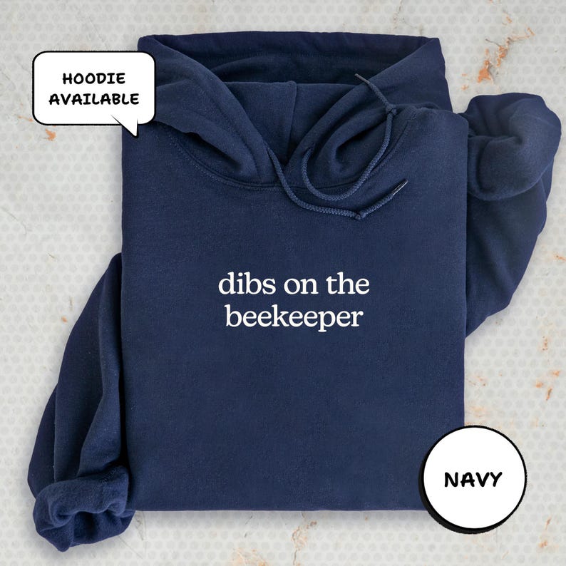 A navy blue hoodie with the text "dibs on the beekeeper" printed on it, along with a speech bubble indicating that the hoodie is available.