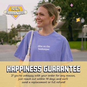 A smiling woman wearing a purple t-shirt with the text "dibs on the beekeeper" is shown in the image.