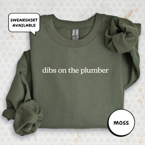 A green sweatshirt with the text "dibs on the plumber" printed on it, along with a speech bubble indicating that the sweatshirt is available.