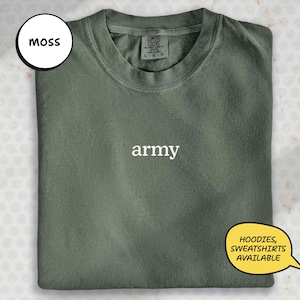 May include: A moss green sweatshirt with the word "army" in white text. The shirt is folded and has a tag that says "Comfort Colors". A speech bubble says "Hoodies, Sweatshirts Available".