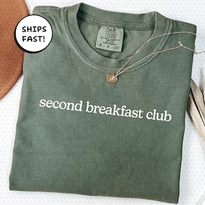 Op de afbeelding: Een olijfgroen t-shirt met de witte tekst "second breakfast club". Een delicate zilveren ketting met een kleine hanger ligt op het shirt. Het shirt heeft een Comfort Colors label.