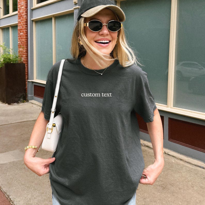 A smiling woman wearing a gray t-shirt with the text "custom text" and sunglasses stands in front of a building.