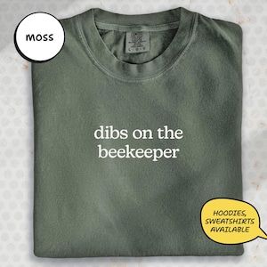 A green t-shirt with the text "dibs on the beekeeper" printed on it, along with a label on the collar.