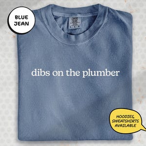 A blue t-shirt with the text "dibs on the plumber" printed on it, along with a label indicating it is a "DIBS ON THE PLUMBERS".