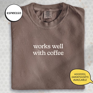 May include: A folded, espresso-coloured sweatshirt with the text "works well with coffee" in white. The sweatshirt has a crew neck and a tag that reads "Comfort Colors". A speech bubble says "Hoodies, Sweatshirts Available".
