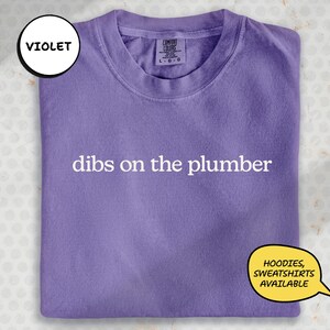 A purple t-shirt with the text "dibs on the plumber" printed on it, along with a label indicating it is a "VIOLET" item.