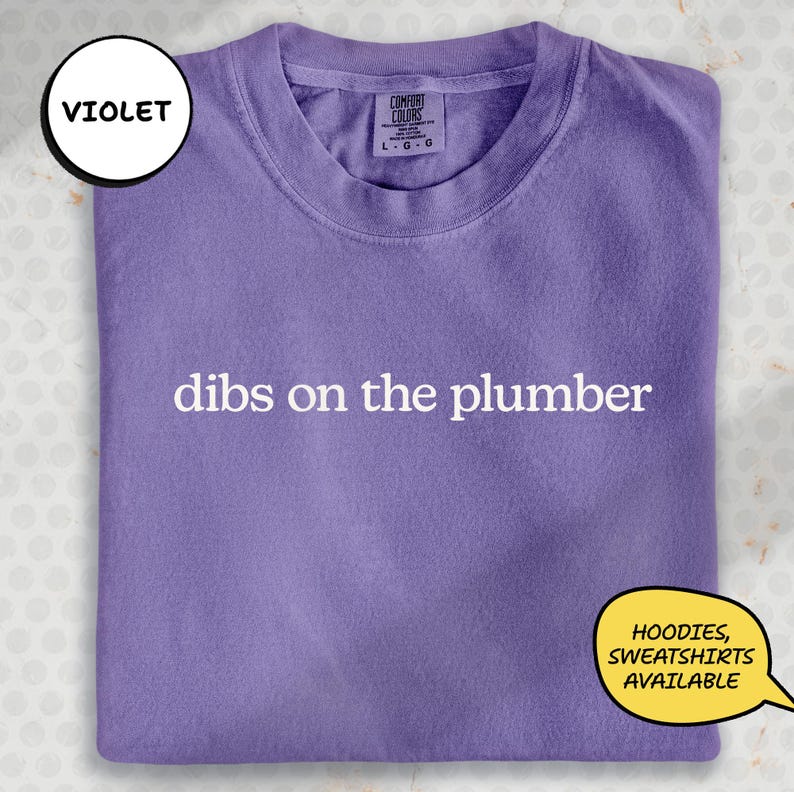 A purple t-shirt with the text "dibs on the plumber" printed on it, along with a label indicating it is a "VIOLET" item.