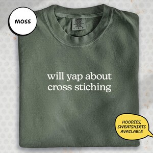 May include: A moss green t-shirt with the text "will yap about cross stitching" in white. The shirt has a crew neck and a tag that says "Comfort Colors". A speech bubble says "Hoodies, Sweatshirts Available". A white circle says "Moss".